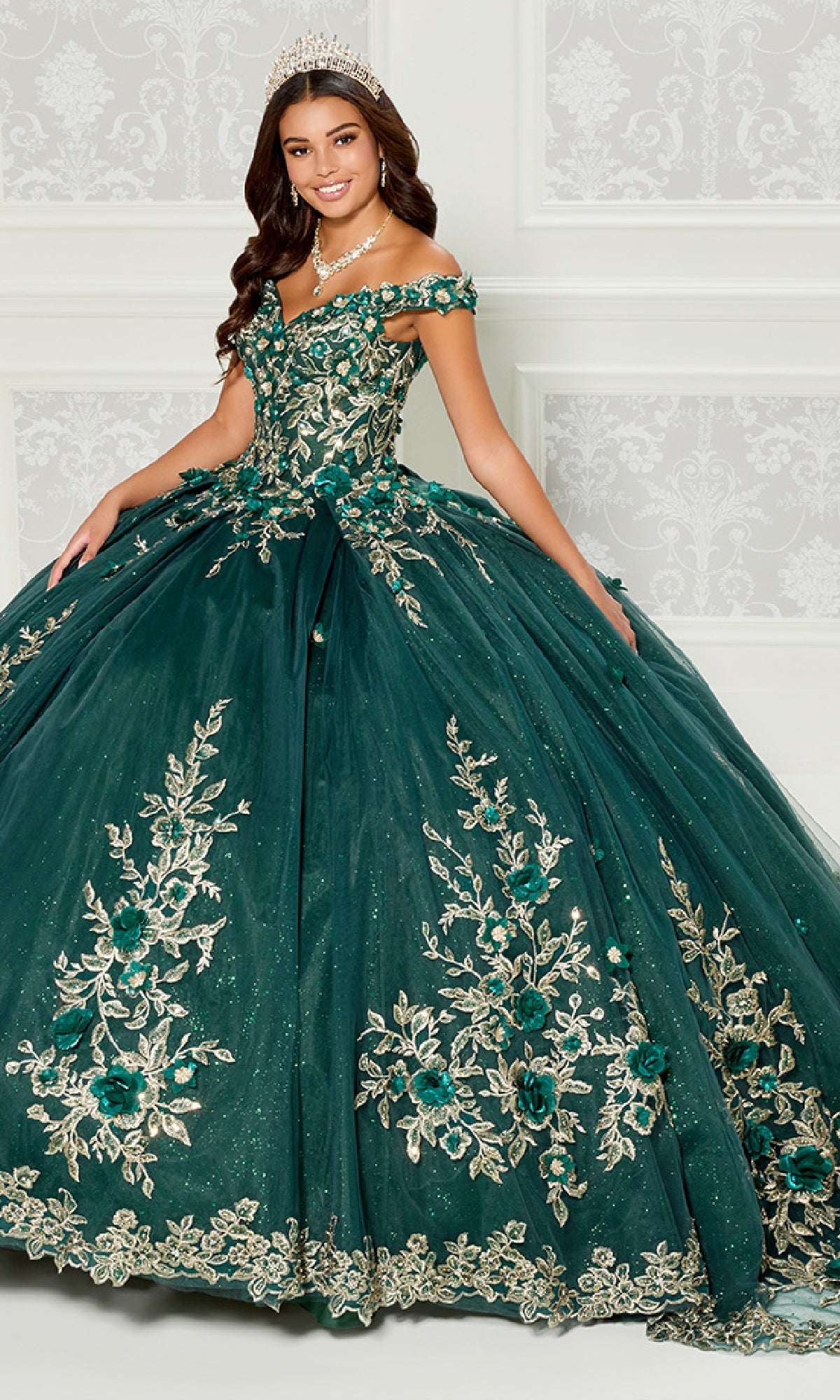 A young woman smiles in an ornate room, wearing the Quinceanera Dress PR30114 by Princesa—an off-the-shoulder ball gown with gold 3D floral appliques and a tiara, her green dress enhancing the regal ambiance.